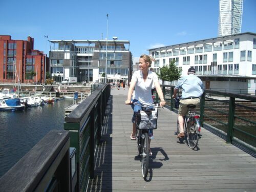 Kvinna cyklar på träbro vid hamn med båtar och moderna byggnader; en man cyklar åt andra hållet.