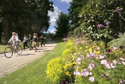 En kvinna cyklar förbi grupp gående ungdomar längs gångstig, färgglada blommor i förgrunden och ett torn i bakgrunden.