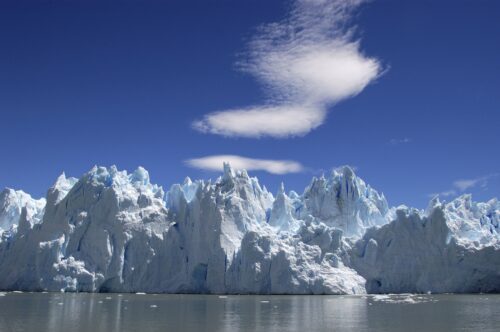 Massiv blåvit patagonisk glaciärfront med spetsiga isformationer som reser sig över stilla vatten under klar blå himmel.
