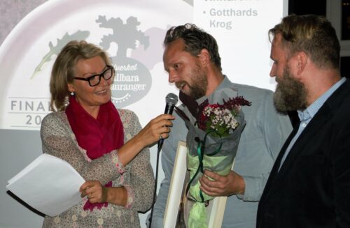 Charlotte Permell håller mikrofonen åt en man som håller blommor och ett pris, medan en annan man tittar på.