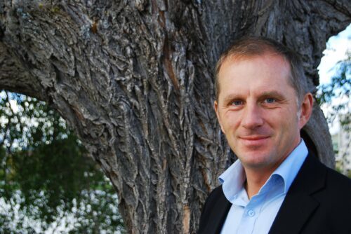 Håkan Wirten, står leende framför grov trädstam, ljusblå skjorta och mörk kavaj.