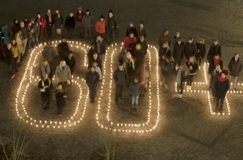 Personer står i och runt en formation av tända ljus som bildar siffrorna "60+"