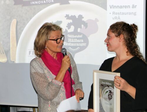 Charlotte Permell till vänster pratar i mikrofon, med Sofia B Olsson till höger som håller ett inramat diplom.
