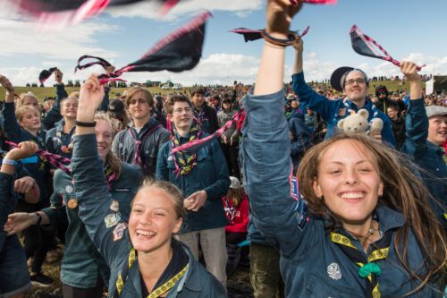 Unga scouter i blå uniformer vinkar med rosa‑svarta halsdukar, ler entusiastiskt.