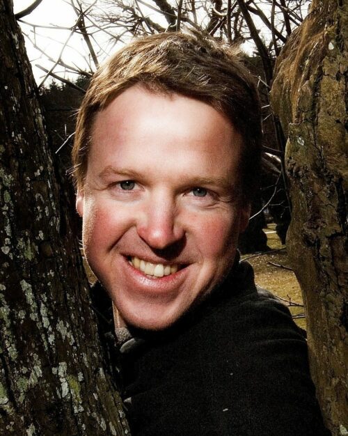 Smilande, Stefan Henningsson med kort ljust hår pressad mellan två trädstammar, ansikte i närbild.