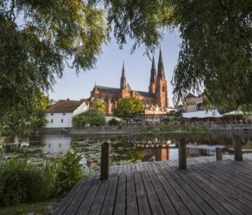 Uppsala domkyrkas röda tegelkyrka med tre höga torn speglas i ån med en träbrygga i förgrunden.