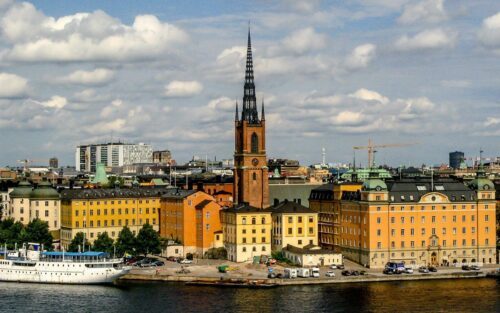 Riddarholmskyrkans spira reser sig över gula byggnader vid vattnet, med en vit passagerarbåt i förgrunden.