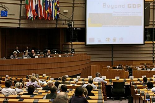 Konferenssal i Europaparlamentet med flera deltagare, panel på podiet och en presentationsskärm med texten "Beyond GDP".
