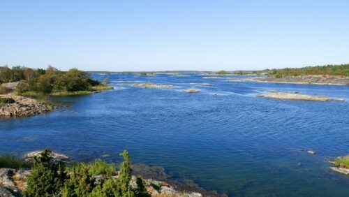 Skärgård med djupblått vatten, små klippöar och gröna buskar under klar blå himmel.