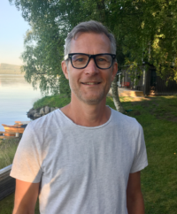 Ola på Ola Bäccman Production AB som står och ler i en vit T-shirt och svarta glasögon framför ett rött hus vid vattnet med björkar på tomten.