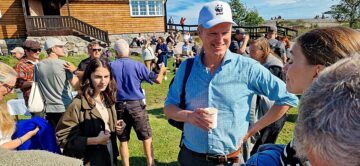 En man i blå skjorta och vit keps med WWFs logga som föreställer en panda håller en kaffekopp utomhus vid ett hus, omgiven av människor.