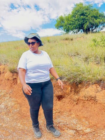 Kvinna i vit t-shirt, en grön hatt och solglasögon går på en solig grusväg kantad av gräs och träd.