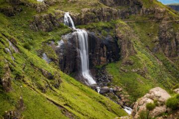 Vattenfall som rinner nerför en klippa omgiven av grönskande gräs och stenar.
