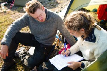 Två personer, en man och en kvinna, sitter bredvid varandra medan kvinnan skriver något på ett vitt papper.