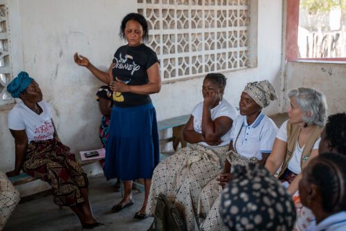 En kvinna står och pratar framför en grupp kvinnor som sitter på bänkar i ett rum med vitmålade väggar.