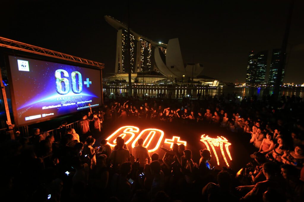 Offentligt Earth Hour firande i Singapore 2014. En stor grupp människor är samlade i en cirkel med ljus i mitten. Ljusen är utplacerade som 60+ och Spiderman-loggan.