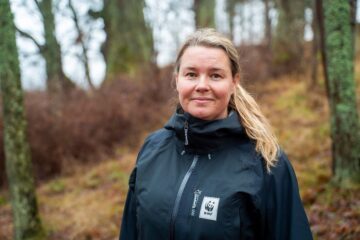 Porträttfoto av Carolina Brändholm, klimatexpert WWF. I bakgrunden syns skog, men ingen grönska och gult gräs. Det ser ut att vara kallt ute.