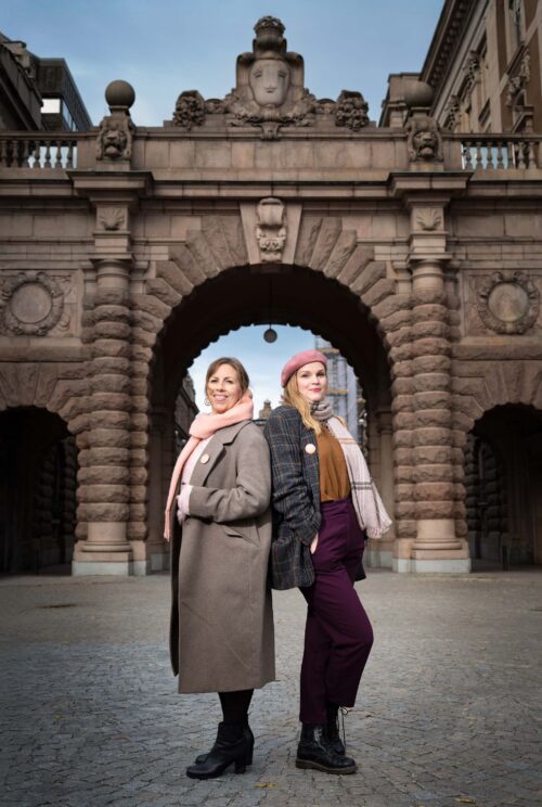 Maria Soxbo och Emma Sundh, grundare av Klimatklubben. De står rygg mot rygg, med byggnaden till Sveriges riksdag bakom sig.