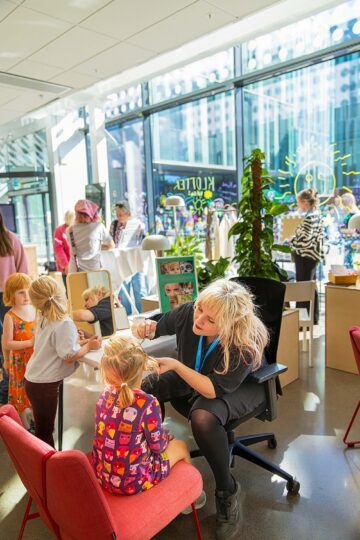 Blond kvinna målar en flickas ansikte, flera barn står i kö i bakgrunden, redo att få ansiktsmålning.