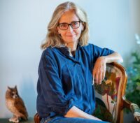 Porträttbild på Helena Nobel, CEO på Wildlife Garden AB. Hon har glasögon och sitter på en stoppad stol i ett ljust rum, klädd i blå skjorta och jeans. I bakgrunden syns gröna växter, en dekorativ uggla och staplade böcker.